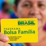 Mão em primeiro plano segurando o cartão amarelo do Programa Bolsa Família do Governo Federal, com uma mulher sorrindo desfocada ao fundo.