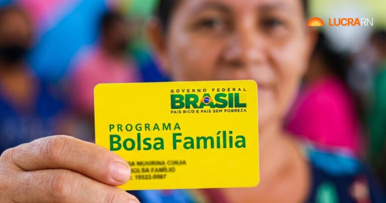 Mão em primeiro plano segurando o cartão amarelo do Programa Bolsa Família do Governo Federal, com uma mulher sorrindo desfocada ao fundo.